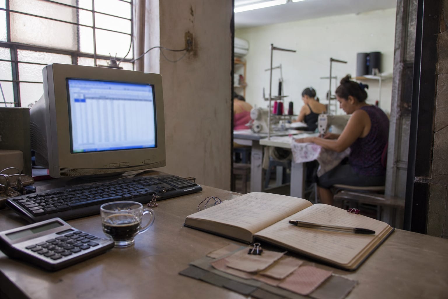 Escritório de facção com caderno de anotações, calculadora e computador, com o salão de costura ao fundo