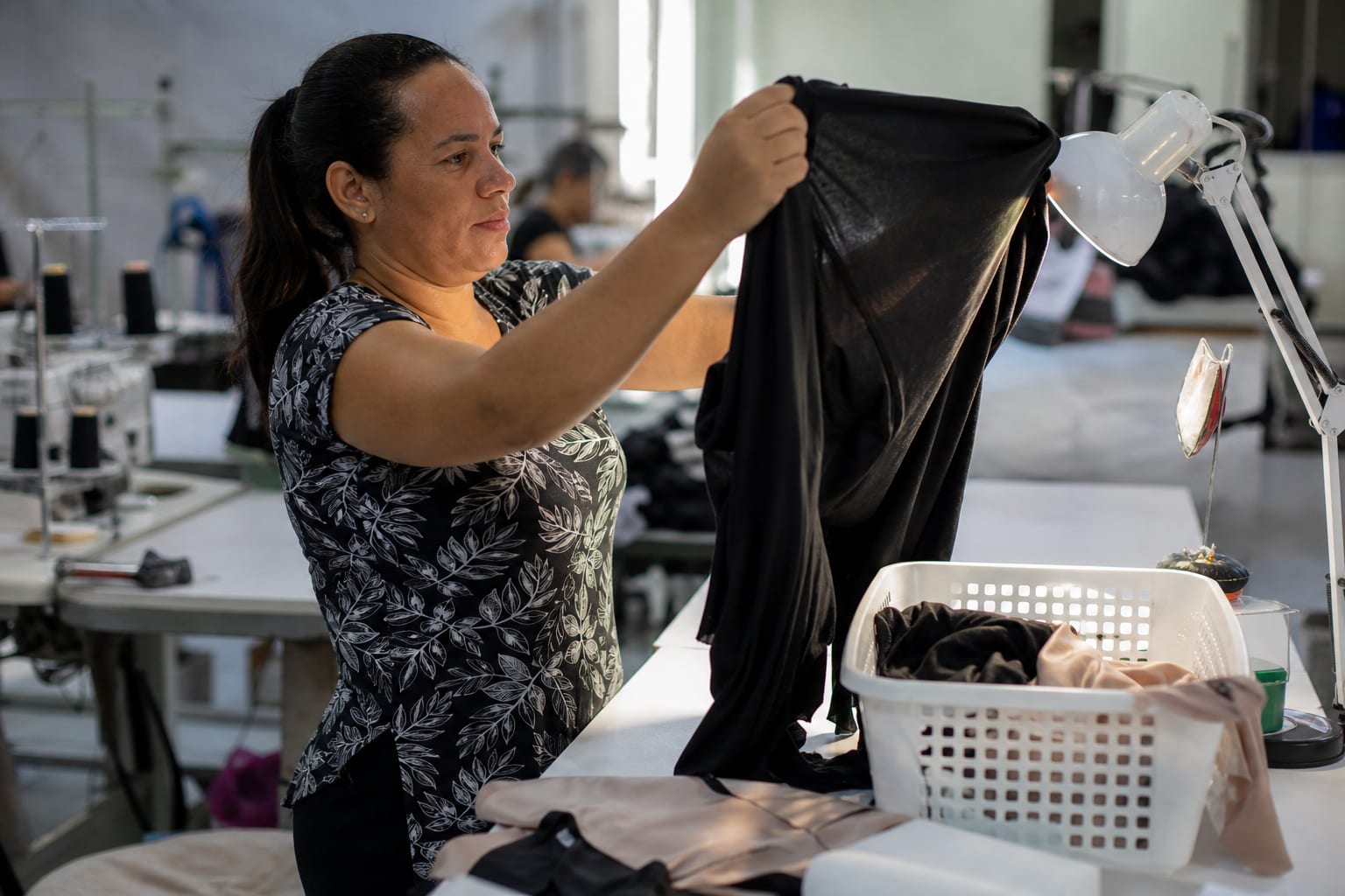 Revisora conferindo uma peça de roupa contra a luz na bancada, com cesto de peças para conserto ao lado